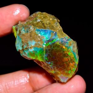 Könnte beinhalten: Ein roher, unpolierter Opal-Edelstein mit einer texturierten, erdbraunen Oberfläche und lebhaften Blitzen aus irisierendem grünem, blauem und orangefarbenem Licht. Der Stein zeigt eine natürliche, unregelmäßige Form mit einem Wabenmuster.
