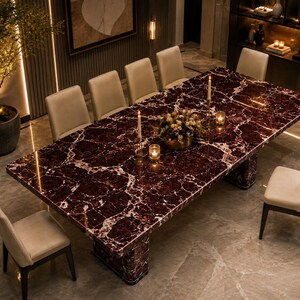 May include: A large rectangular dining table with a deep red and white marble top and two matching pedestal bases, surrounded by eight upholstered chairs with light beige fabric seats and dark wooden legs, arranged in a formal dining room setting.