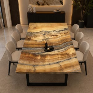 Peut inclure: Une table à manger rectangulaire avec un plateau en pierre d'onyx ambre et brun poli présentant des veines naturelles, soutenue par un cadre en métal noir. Huit chaises rembourrées couleur crème à dossier incurvé entourent la table.