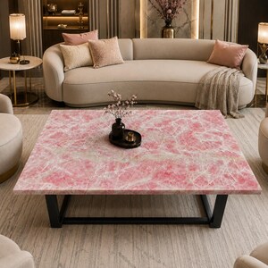 May include: A square coffee table featuring a polished pink marble top with white veining and a black metal frame. The table surface holds a small black vase containing delicate pink floral branches and a small black circular tray.
