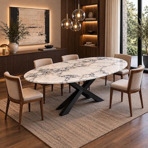 Op de afbeelding: Een ovale eettafel met een wit en grijs marmeren blad met opvallende aders, ondersteund door een zwart metalen X-vormig onderstel. De tafel is omringd door vier stoelen met lichtbeige gestoffeerde zittingen en donkerhouten frames.