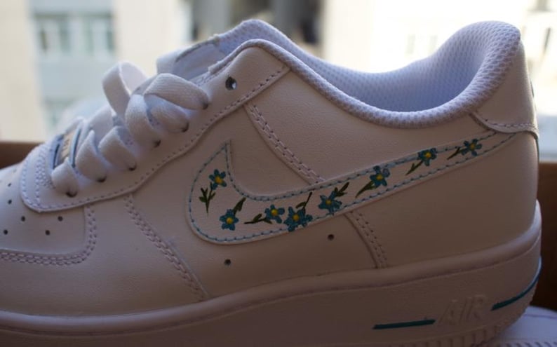 K&ouml;nnte beinhalten: Wei&szlig;e Ledersneaker mit niedrigem Schaft und einem blauen und wei&szlig;en Blumendesign an der Seite. Die Sneaker haben eine wei&szlig;e Sohle mit einem blauen Rand.