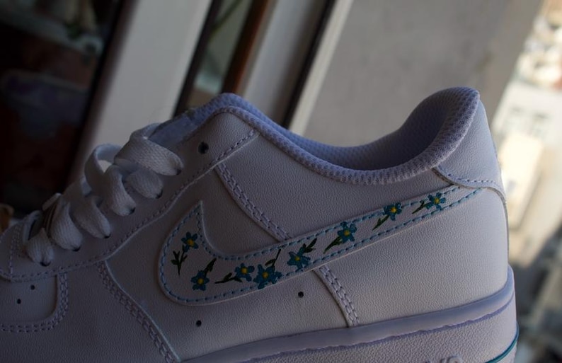 K&ouml;nnte beinhalten: Wei&szlig;e Lederschuhe mit einem blauen und gr&uuml;nen Blumendesign auf dem Swoosh.