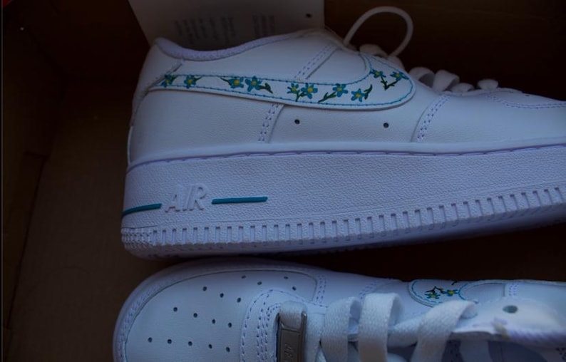 K&ouml;nnte beinhalten: Wei&szlig;e, niedrige Lederschuhe mit einem blauen und gr&uuml;nen Blumendesign an der Seite. Die Schuhe haben eine wei&szlig;e Sohle mit dem Wort "AIR" in Blau.