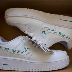 Close-up of the hand-painted floral design on white Nike Air Force 1 sneakers, showcasing vibrant blue and green detailing.