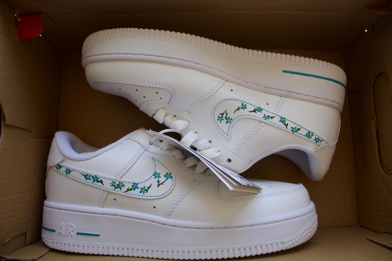 Close-up of the hand-painted floral design on white Nike Air Force 1 sneakers, showcasing vibrant blue and green detailing.