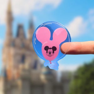 May include: A clear sticker featuring a pink Mickey Mouse balloon design within a blue bubble. The Mickey Mouse balloon has a smiling face and is held up by a finger against a blurred background of a castle and blue sky.