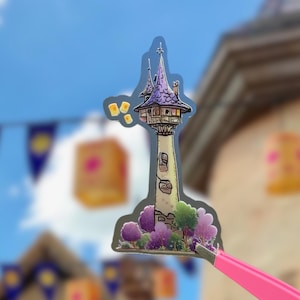 May include: A sticker featuring a cartoon illustration of a tall tower with a purple roof and a small balcony. The tower is surrounded by purple and green trees. Floating lanterns are in the background. The sticker is held by pink tweezers.