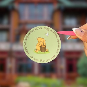May include: A round, light green sticker featuring Winnie the Pooh with a honey pot. The sticker has the text "You're braver than you believe, stronger than you seem, and smarter than you think."