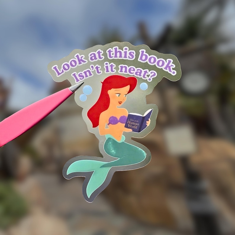 May include: A sticker featuring a cartoon mermaid with red hair, a purple top, and a teal tail, reading a book. The sticker has the text "Look at this book, Isn't it neat?" and is held by pink tweezers.