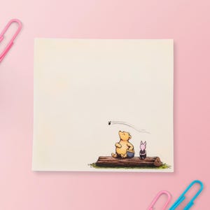 May include: A square notepad featuring a printed illustration of a yellow bear and a small pink pig sitting on a brown log. The bear holds a blue honey pot while looking up at a small black bee flying in the air. The paper is off-white.