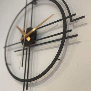 May include: A contemporary wall clock with a black metal frame and gold hands. The clock has a circular design with linear details, creating a geometric look. The clock's hands show the time. The clock is mounted on a white wall.