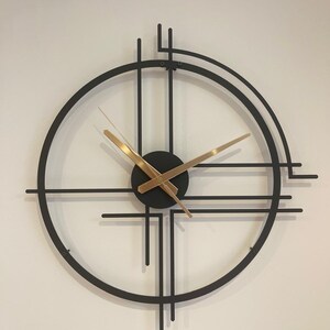 May include: A contemporary wall clock with a black metal frame and gold hands. The clock has a circular design with intersecting lines and a small black centre. The hands are thin and elegant, showing the time.