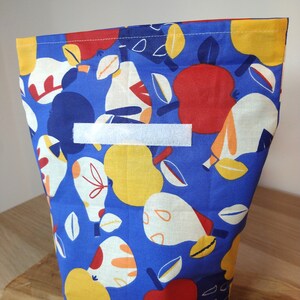 May include: A blue fabric reusable bag with a white label and a pattern of red apples, yellow pears, and white leaves.