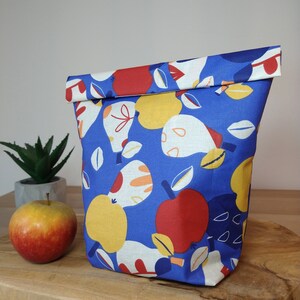 May include: A blue fabric lunch bag with a colorful pattern of apples and pears. The bag is folded over at the top and has a white lining.