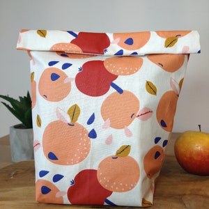 May include: A white fabric lunch bag with a repeating pattern of orange and red apples with blue and yellow accents. The bag is folded over at the top and has a white background.