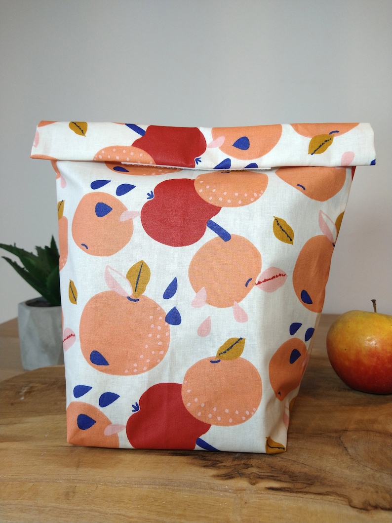May include: A white fabric lunch bag with a repeating pattern of orange and red apples with blue and yellow accents. The bag is folded over at the top and has a white background.