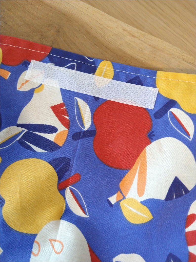 May include: A blue fabric with a white, red, and yellow fruit pattern. A white hook and loop fastener is sewn onto the fabric.