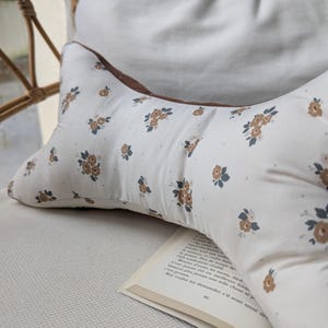 May include: A floral-patterned pillow with a brown trim and a white background. The pillow is resting on a light-colored surface, with a book open in front of it. The pillow has a pattern of small brown and blue flowers.