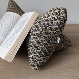 Op de afbeelding: Een leeskussen met een beige en marineblauw golfpatroon ondersteunt een open boek. Het wigvormige kussen is ontworpen om boeken te ondersteunen voor handsfree lezen. Het boek is open op een pagina met tekst.