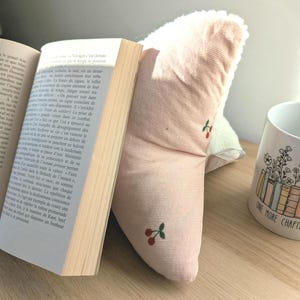 May include: A pink book pillow with a cherry pattern supports an open book. A white mug with the text "ONE MORE CHAPTER" and a floral design sits nearby. The pillow has a white trim.