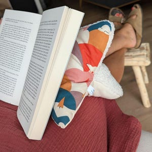 May include: A book is propped open by a patterned pillow with a white, textured border. The pillow features a design with orange, teal, and pink shapes. The person is wearing a red shirt and brown sandals.