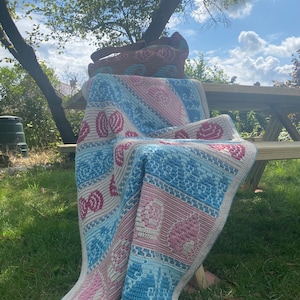 May include: A blue and pink crocheted blanket with a geometric pattern is draped over a wooden bench. The blanket is made with a variety of stitches and colors, creating a unique and eye-catching design.