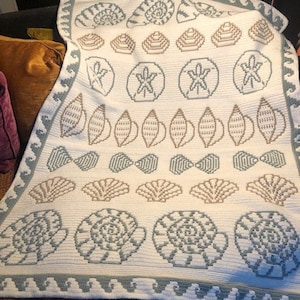 Pode incluir: Uma manta de crochê branca com um design de conchas azuis e marrons. A manta apresenta um padrão repetitivo de conchas, estrelas do mar e ondas.