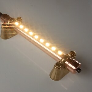 May include: A linear light fixture featuring a copper-toned cylindrical tube with a row of small, illuminated LED bulbs. The tube is supported by two brass-colored metal mounts and is connected to a black electrical power cord with a plug.