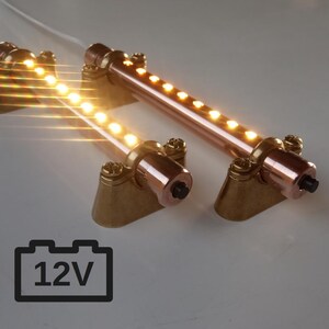 Könnte beinhalten: Zwei kupferfarbene LED-Lichtleisten mit warmweißen Lichtern. Jede Leiste ist auf einer messingfarbenen Basis montiert und mit Schrauben befestigt. Das Bild enthält ein 12V-Batteriesymbol.