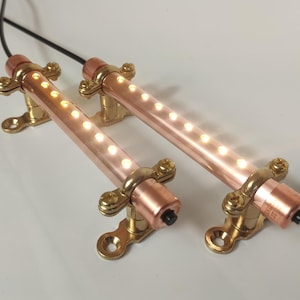 May include: Two copper pipe light fixtures with brass fittings and warm white LED lights. The fixtures are mounted on a white surface.