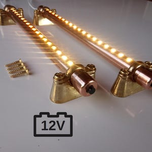 Könnte beinhalten: Zwei LED-Lichtleisten aus Kupfer und Messing mit warmweißen Lichtern. Die Leisten haben eine Reihe kleiner Lichter und sind auf Messingbasen montiert. Das Bild zeigt auch einen Satz Schrauben und ein 12V-Batteriesymbol.