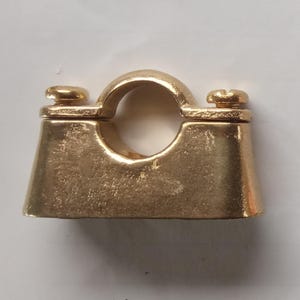 Puede incluir: Una abrazadera de tubería de color dorado con una parte superior redondeada y dos tornillos. La abrazadera tiene una abertura circular en el centro y una base trapezoidal. El metal tiene una superficie ligeramente texturizada.