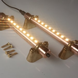 May include: Two copper-colored LED light fixtures with brass mounting brackets and a row of white LED lights. The fixtures are on a white surface with four gold screws.
