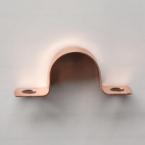 Könnte beinhalten: Eine kupferfarbene Rohrschelle mit einem gebogenen, U-förmigen Design und zwei flachen Enden. Jedes Ende hat ein kreisförmiges Loch zur Montage. Die Schelle besteht aus glänzendem Metall.