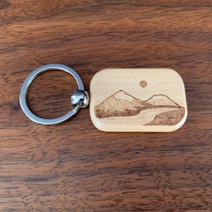 May include: Wooden keychain with a laser-engraved image of a mountain range and a sun. The keychain is attached to a silver metal ring.