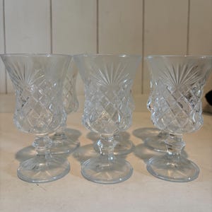 May include: A set of clear, vintage-style crystal goblets. Each goblet features a diamond-cut pattern on the bowl and a detailed stem. The glasses are arranged on a white surface, showcasing their elegant design.