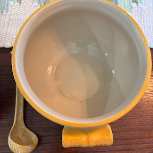 Puede incluir: Un cuenco de cerámica con exterior amarillo e interior crema. El cuenco tiene una base amarilla decorativa y un cucharón a juego con un patrón moteado. El cuenco está diseñado para servir o exhibir.