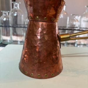 May include: A copper-colored Turkish coffee pot with a hammered texture. The pot has a flared top, a brass handle, and is adorned with small brass rivets around the rim and base. The pot is designed for brewing coffee.