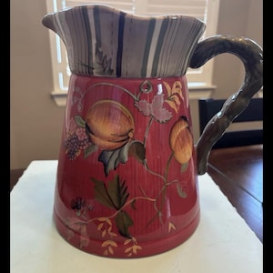 May include: A decorative ceramic pitcher with a red base and a brown handle. The pitcher features a fruit and leaf design in shades of orange, green, and purple. The top rim has a striped pattern in gray, brown, and green.