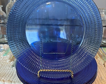 May include: A stack of circular cobalt blue glass plates featuring a textured rim with a repeating vertical ribbed pattern. The plates are displayed upright on a gold-toned metal stand, showcasing the translucent quality and deep blue color.