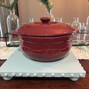 May include: A burgundy ceramic casserole dish with a lid, set on a light blue wooden tray. The dish has handles and a textured design. The tray has white, rounded feet and decorative studs. The background includes glass vases and floral arrangements.