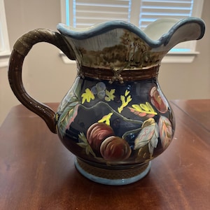 Puede incluir: Una jarra decorativa de cerámica con base azul oscuro, con diseños botánicos y paisajísticos pintados a mano. La jarra tiene un asa marrón y un borde azul claro. La obra de arte incluye hojas, bayas y una escena de bosque. La jarra mide unos 25 cm de altura.