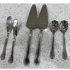 Könnte beinhalten: Ein Set aus sechs silberfarbenen Edelstahlbestecken, bestehend aus zwei Buttermessern mit abgerundeten Spitzen, zwei dreieckigen Tortenhebern mit flachen Klingen und zwei kleinen Löffeln mit gewelltem Rand und verzierten Griffen.