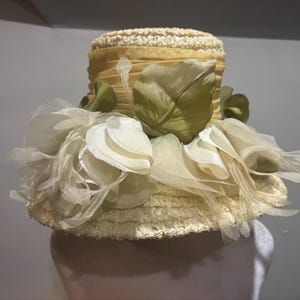 May include: A vintage straw hat with a wide brim and a cylindrical crown. The hat is adorned with cream-colored fabric petals and green silk leaves. The hat has a yellow ribbon around the crown.