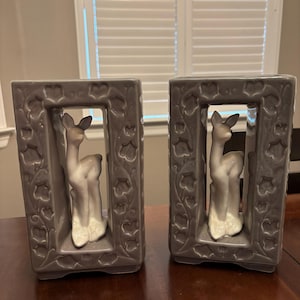 Puede incluir: Dos figuras de ciervos de cerámica gris a juego dentro de marcos rectangulares. Los marcos tienen un diseño floral en los bordes. Los ciervos son blancos y grises con detalles. Las figuras se exhiben sobre una mesa de madera.