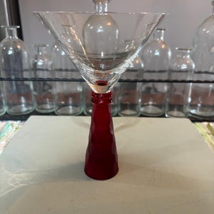 May include: A martini glass featuring a wide, clear, triangular bowl and a solid, deep red stem with a faceted, geometric texture. The base of the stem is circular, providing stability to the glass, which stands upright on a flat surface.