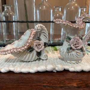 May include: A white ceramic decorative set featuring a small pitcher and a shell-shaped basket. Both pieces have pink ruffled edges, gold accents, and a pink rose with green leaves. The set is displayed on a white textured surface.