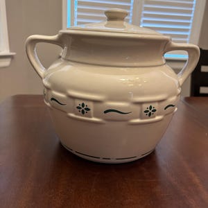 Pode incluir: Pote de cerâmica creme com tampa e duas alças. O pote apresenta uma faixa decorativa com detalhes florais e curvos verdes. O pote está sobre uma mesa de madeira marrom escuro.