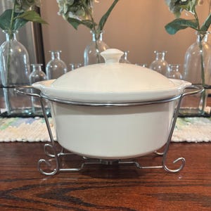 May include: A white ceramic casserole dish with a lid, set on a chrome-colored metal stand. The dish has a rounded shape and a small handle on the lid. The stand has a decorative, curved design.
